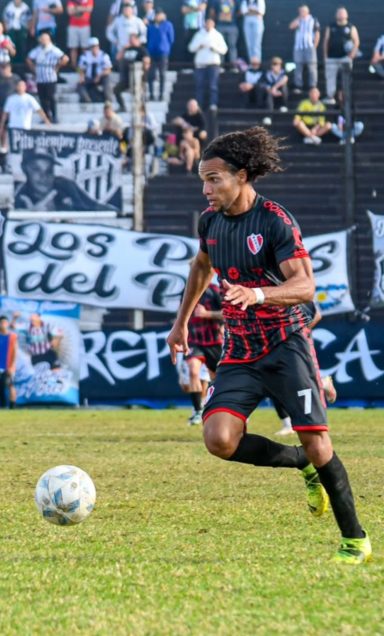 Jugador de fútbol corriendo con balón en un campo, aficionados en las gradas.